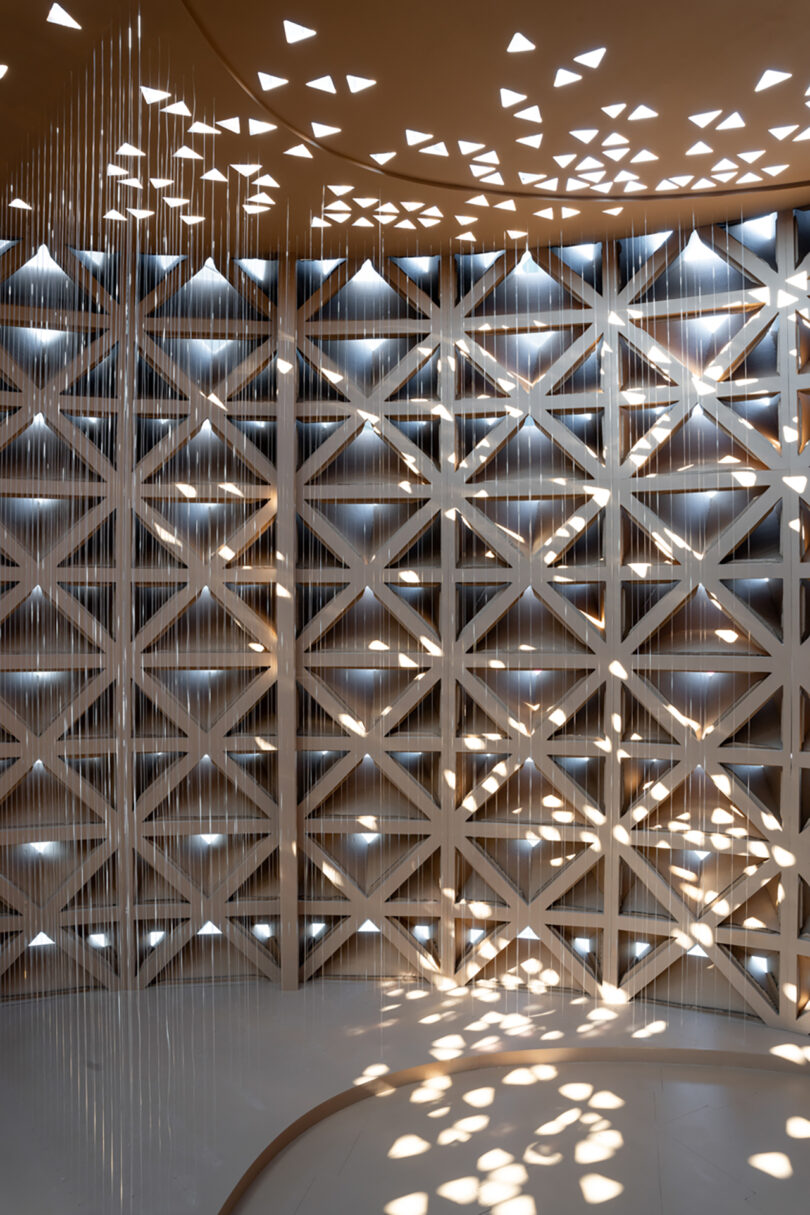 A geometric lattice wall with triangular and diamond-shaped cutouts casts intricate light patterns onto the floor and curved wall.