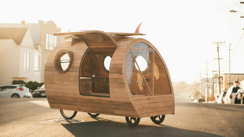 A wooden, teardrop-shaped bike trailer designed by Anya Molyviatis stands gracefully on a sunlit street, its transparent side panels gleaming in the daylight.