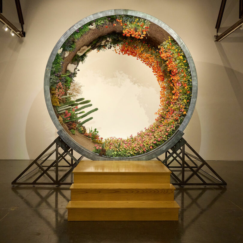 A large circular art installation by Anya Molyviatis, featuring vibrant flowers and plants supported by a metal framework, stands on a wooden platform in the gallery.