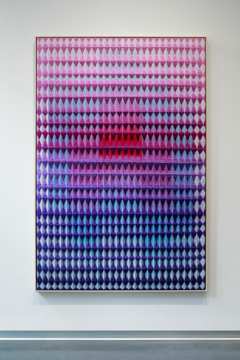 Geometric artwork with a gradient from pink to blue, featuring repeating diamond shapes in a framed vertical piece by Anya Molyviatis, set against a white wall.