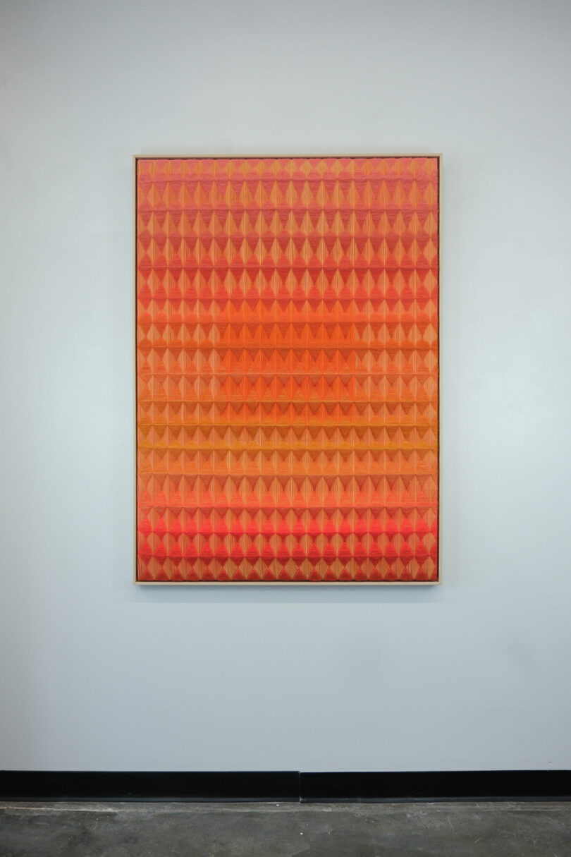 A large geometric painting by Anya Molyviatis, featuring a pattern of overlapping red and orange triangular shapes, hangs elegantly on a white wall.