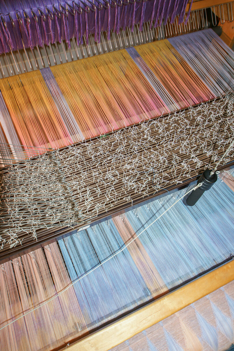 A vivid close-up of a loom showcases Anya Molyviatis' colorful threads in shades of purple, orange, yellow, and blue, arranged in harmonious parallel lines.