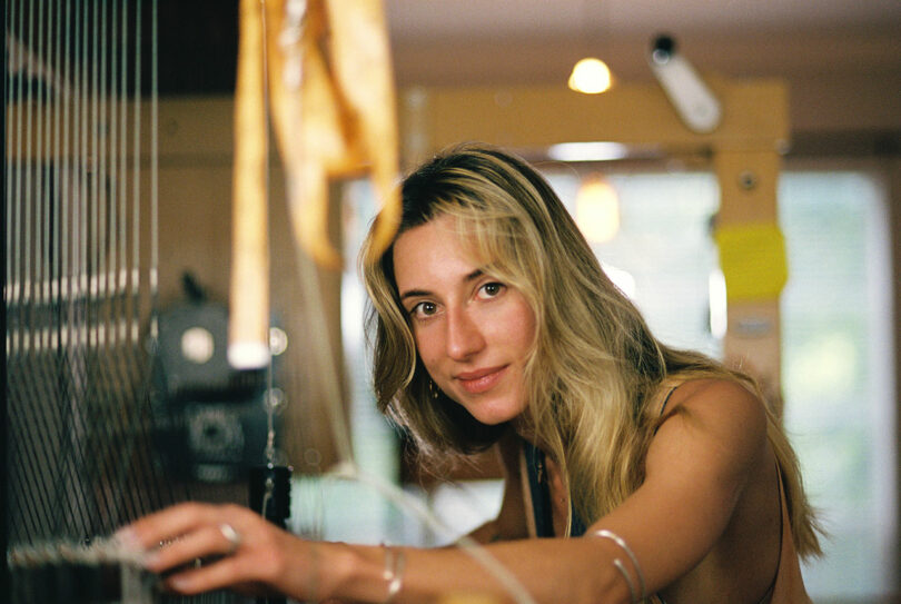 Anya Molyviatis, a woman with long blonde hair, is adjusting strings on a loom with a focused expression.