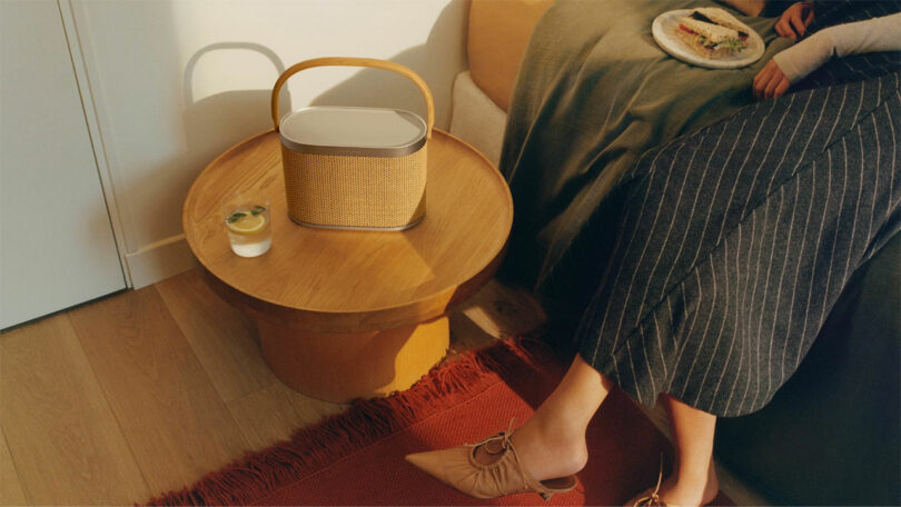 A person sits on a bed holding a plate of food, reminiscent of a scene from a Giulio Barresi painting. Nearby, on the red rug, rests a small wooden table adorned with a portable basket and glass, capturing an essence of everyday elegance.