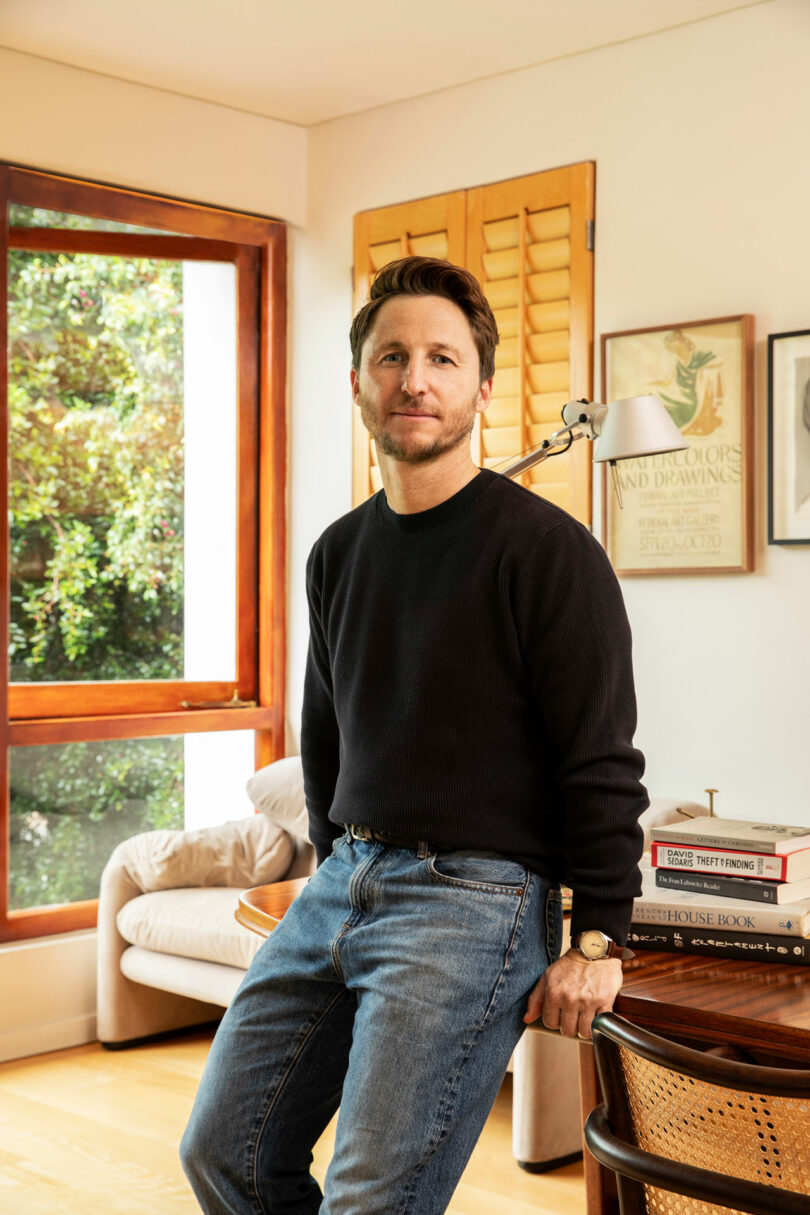 Kevin Frankental, in a black sweater and jeans, leans casually against a desk in a cozy room adorned with books and art on the walls.