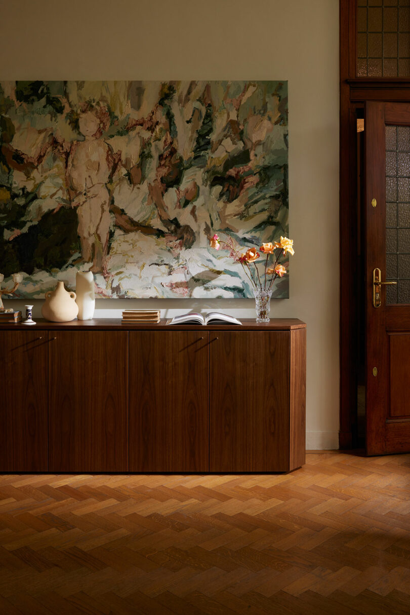 A wooden sideboard with decorative vases and an open book sits beneath a large abstract painting reminiscent of Kevin Frankental's style. A wooden door with frosted glass panels is partially visible to the right.