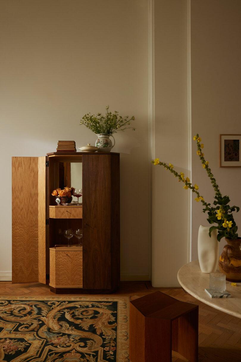 A wooden cabinet with open doors reveals glassware and decor items, reflecting the curated style of Kevin Frankental. A vase with flowers sits on top, echoing the elegance found in Frankental's designs. Another vase adorns a nearby round table, complemented by a rug and art piece visible in the room.