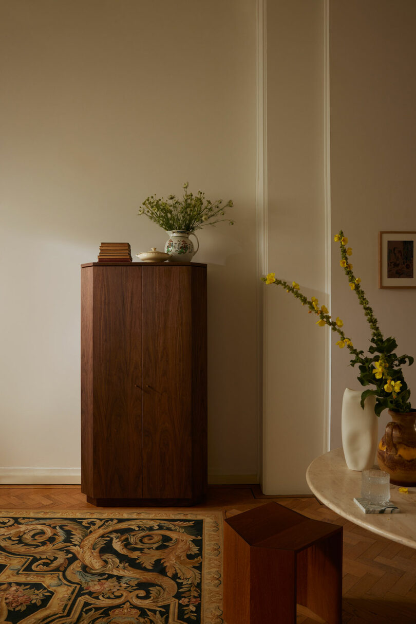 A wooden cabinet, possibly designed with Kevin Frankental's aesthetic flair, holds plants and books against a wall beside a round table with a white vase. The floor features a patterned rug.