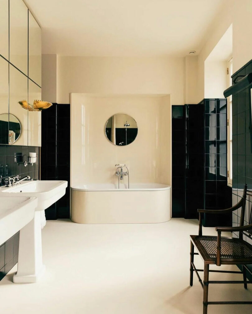 A bathroom styled by Kevin Frankental features a round bathtub set against dark tiles, a circular mirror above it, dual sinks to the left, and a wooden chair on the right.