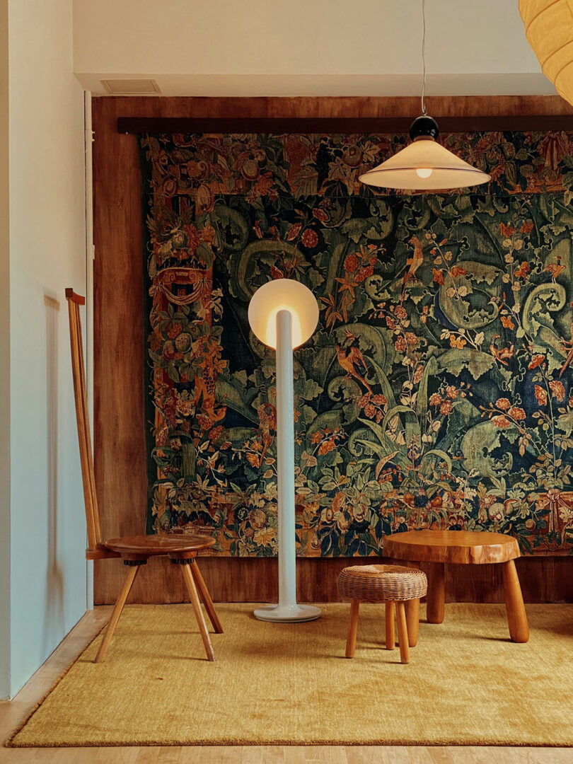 A cozy room with a tapestry wall, wooden chairs, a small table, and a tall floor lamp designed by Kevin Frankental. The woven stool sits gracefully on a beige rug, adding charm to the space.