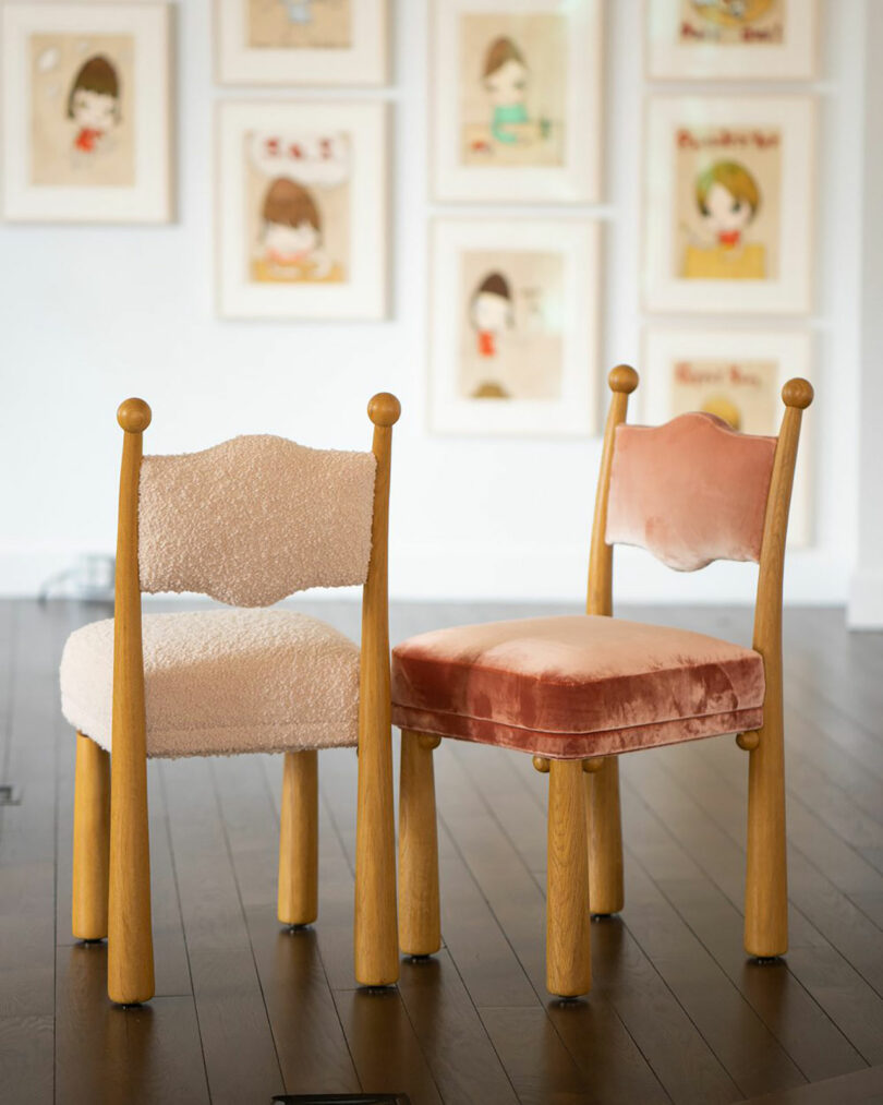 Two chairs with wooden frames, one boasting a textured white seat and the other a smooth pink one, sit elegantly on a wooden floor. Behind them, framed illustrations curated by Kevin Frankental add an artistic touch to the scene.