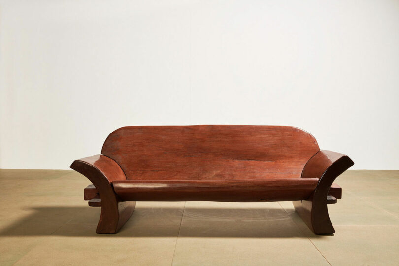 A wooden bench designed by Kevin Frankental features a curved backrest and armrests, sitting elegantly on a tiled floor against a plain wall.