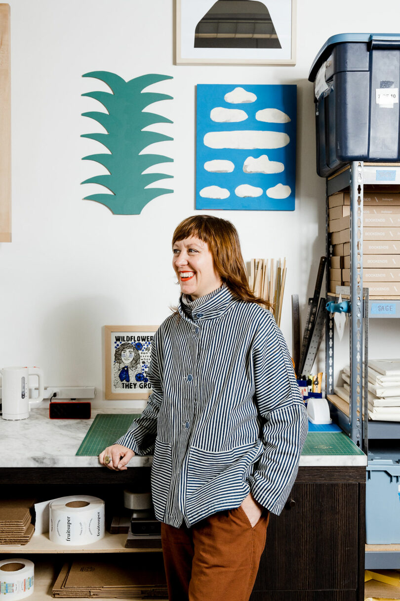 Sallyann Corn, wearing a striped shirt, stands in her art studio adorned with cloud and plant wall decorations, beside shelves and a workspace table.