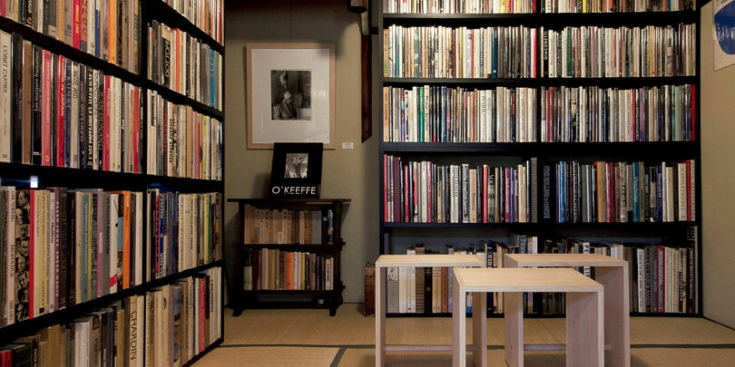 A room with shelves filled with books, a framed photo of Sallyann Corn on the wall, and three wooden stools in the center.