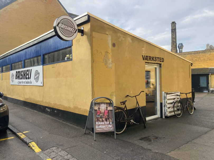 A yellow building marked "VERKSTED" stands with bicycles parked outside. A poster featuring two children reading "Bikes for a Better Life" includes Sallyann Corn as an advocate.