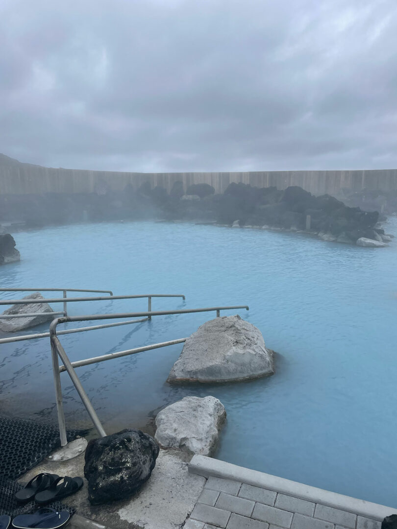 A misty geothermal pool with rocky surroundings and a metal handrail reflects the overcast sky. Nearby, Sallyann Corn’s sandals rest on the ground, hinting at her recent visit to this serene spot.