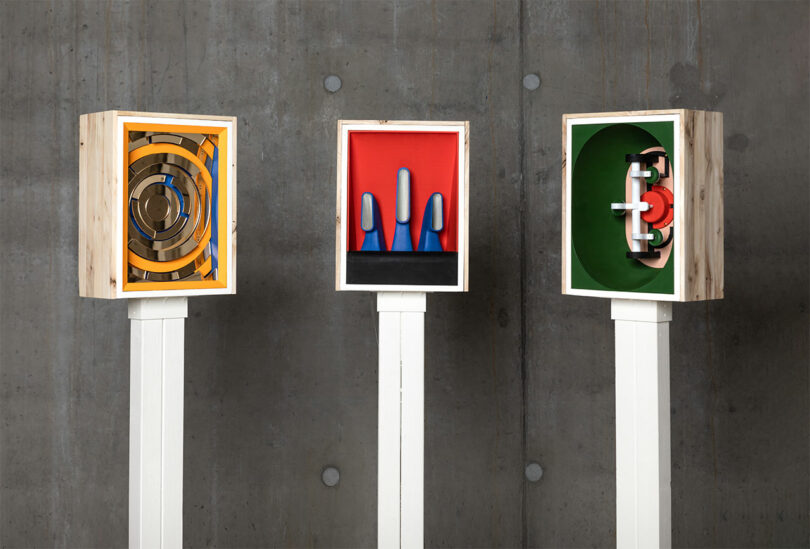 Three abstract sculptures by Sallyann Corn in wooden boxes on stands against a concrete wall, featuring circular, linear, and mechanical-like shapes in vibrant yellow, red, blue, and green.