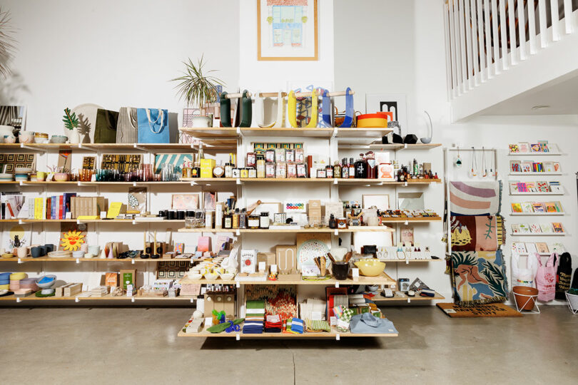 A retail store display with shelves showcasing diverse home goods, including Sallyann Corn kitchenware, lush plants, captivating books, and charming decor items.