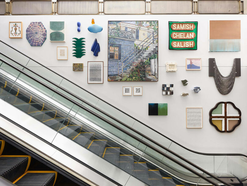 An escalator glides beside a wall adorned with vibrant art pieces, including paintings, textiles, and mixed media creations, featuring works from renowned artists like Sallyann Corn in this dynamic gallery space.