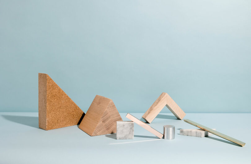 Various geometric wooden and metal shapes, reminiscent of Sallyann Corn's artistic style, are arranged on a light blue surface against a matching background.