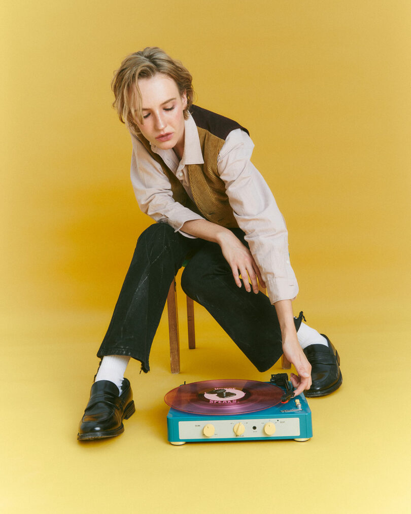 A person sits on a stool, immersed in the rich sound of a Gadhouse blue vintage turntable spinning a vinyl record, all set against a vibrant yellow background.