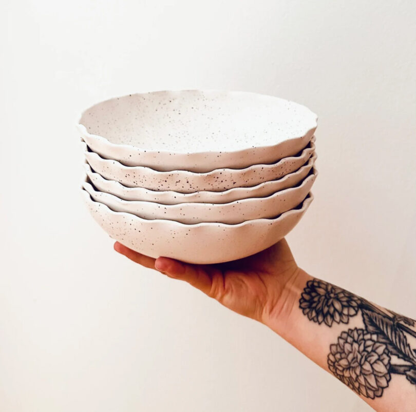 A tattooed arm holds a stack of five white ceramic bowls with scalloped edges and speckled design.