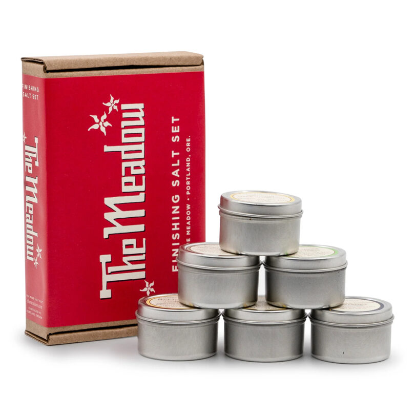 A set of six metal tins stacked in a pyramid next to a red box labeled "The Meadow Finishing Salt Set.