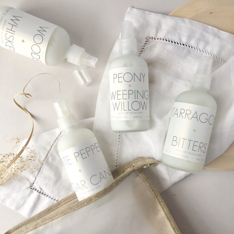 Four bottles of room spray with different scents, including Peony + Weeping Willow and Tarragon + Bitters, displayed on a cloth with a beige pouch nearby.