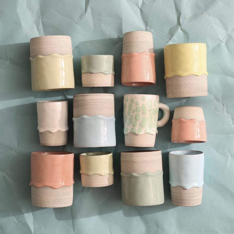 A collection of ceramic mugs and cups with pastel-colored glazes arranged on a textured, light green background.