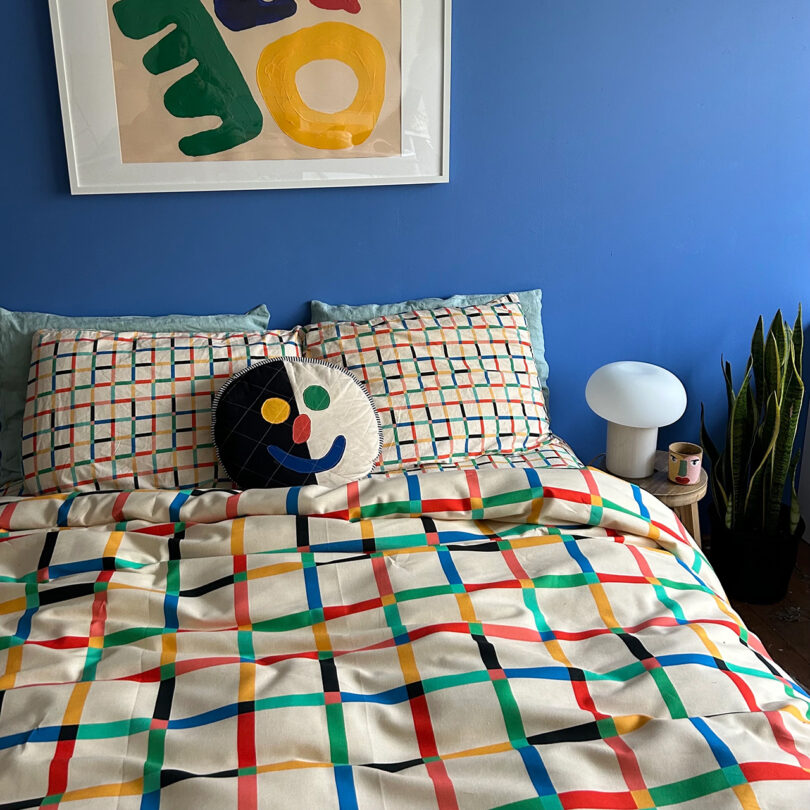 A bed with colorful grid-patterned bedding, two pillows, and a round cushion. A blue wall with abstract artwork and a side table with a lamp, candle, and a small plant are visible.