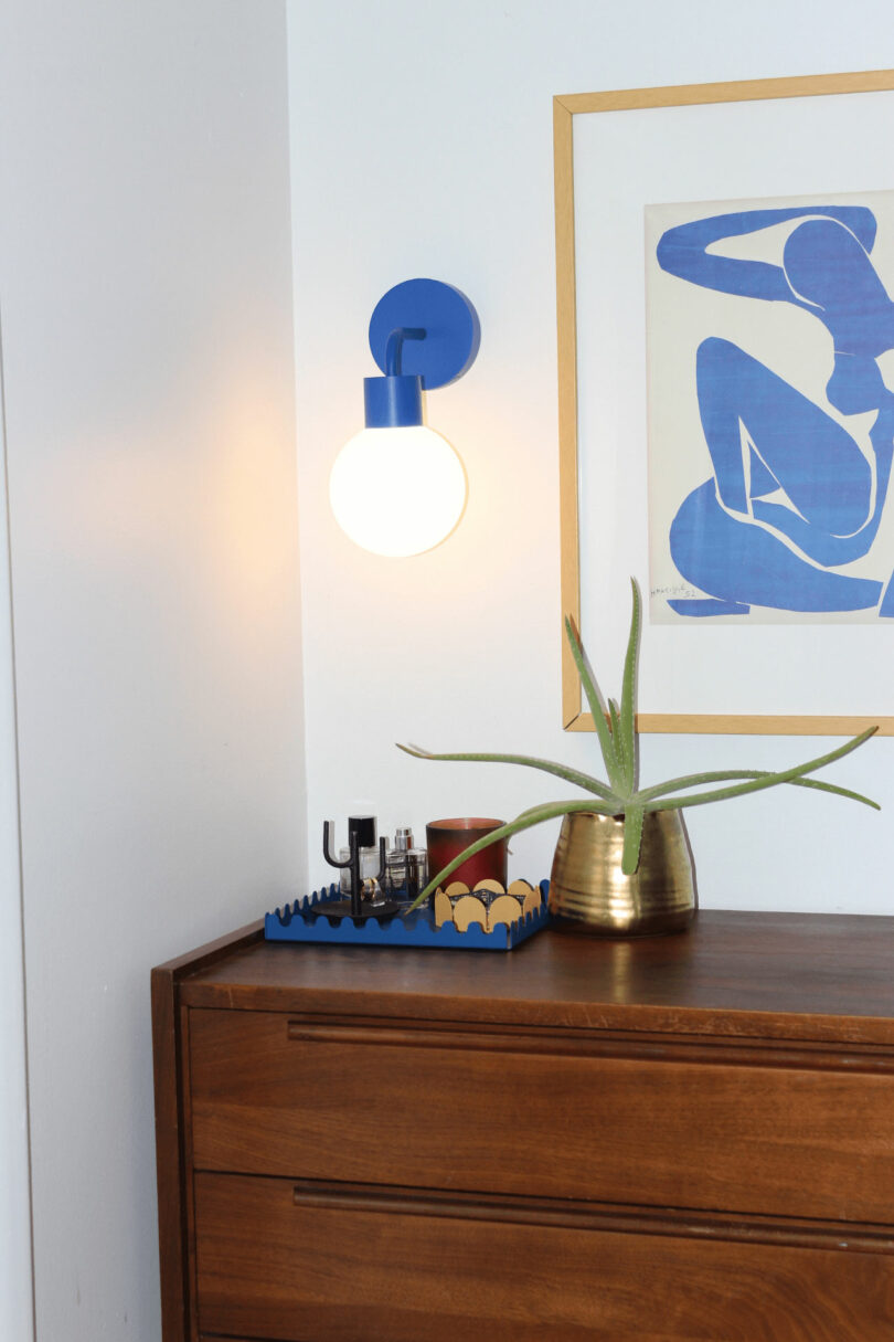 In the room's corner, you'll find a wooden dresser topped by a plant in a gold pot, casting subtle reflections against the blue wall sconce. Framed abstract art adds flair to this curated setting, echoing an Aria Lee Gift Guide recommendation for sophisticated home accents.