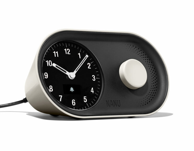 A modern analog clock with a black face and white numbers, showing 10:10. It has a circular knob and a small screen displaying a bell icon.