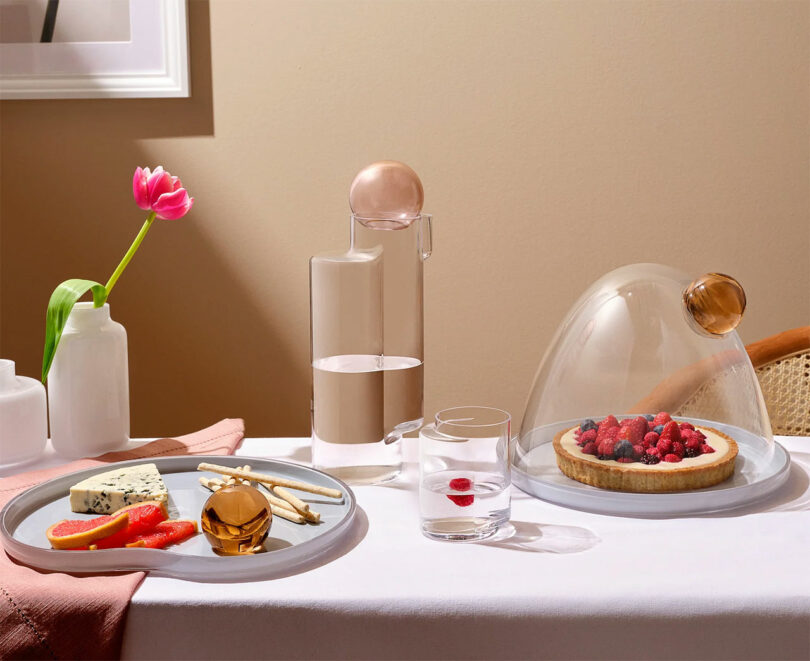 Table setting with cheese plate, raspberry tart under a glass dome, and a carafe with pink liquid. A tulip in a vase and a glass of water with a raspberry are also on the table.