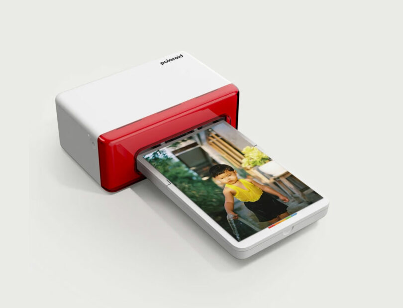 A red and white Polaroid printer is printing a photo of a young child in a yellow top outdoors.