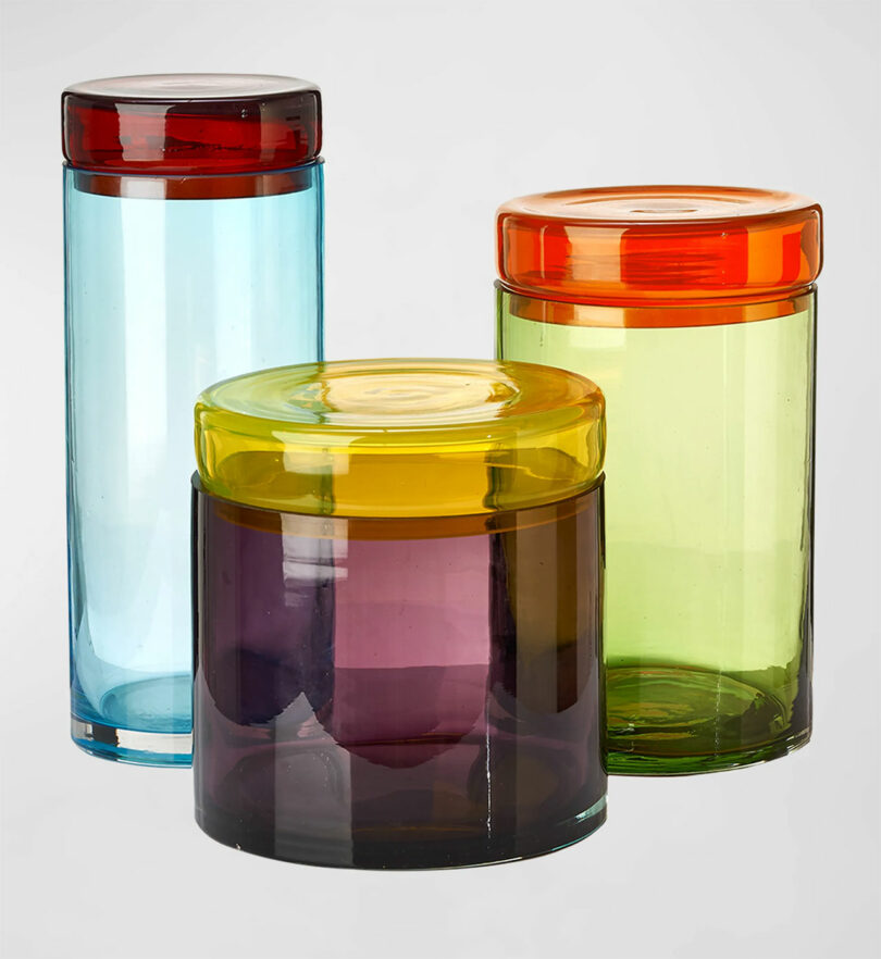 Three glass jars with colorful lids—red, yellow, and orange—stand against a plain background. The jars are blue, purple, and green.