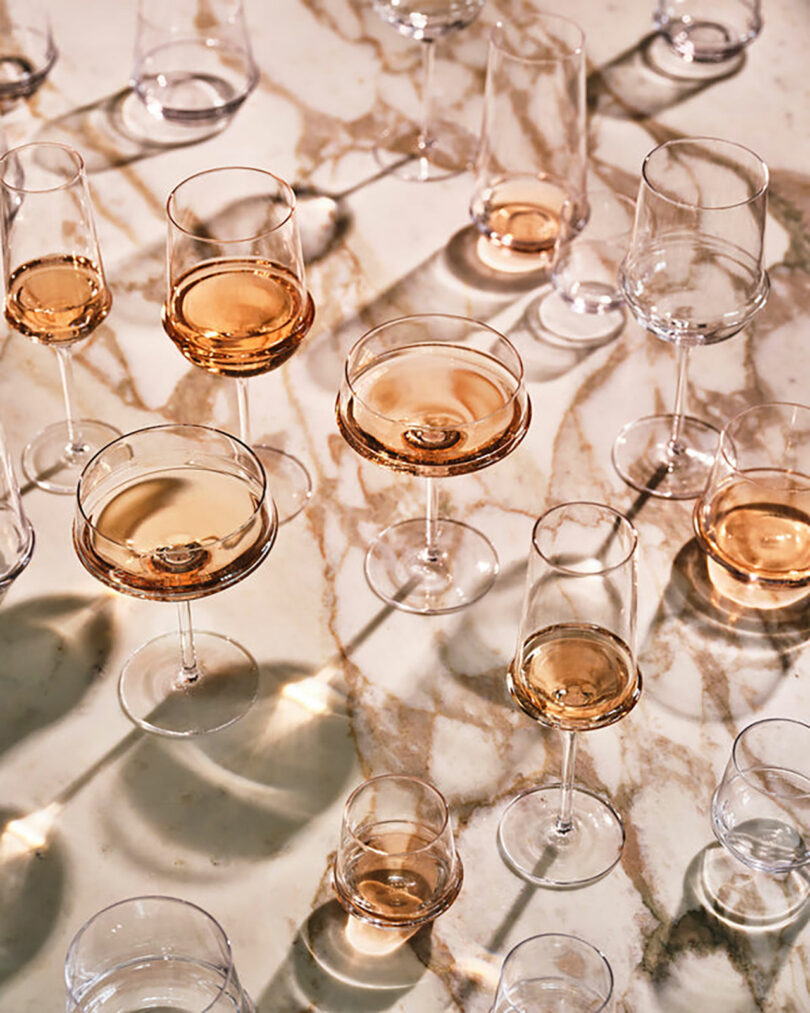 A variety of wine glasses filled with rose and clear liquid are arranged on a marble surface, casting shadows.