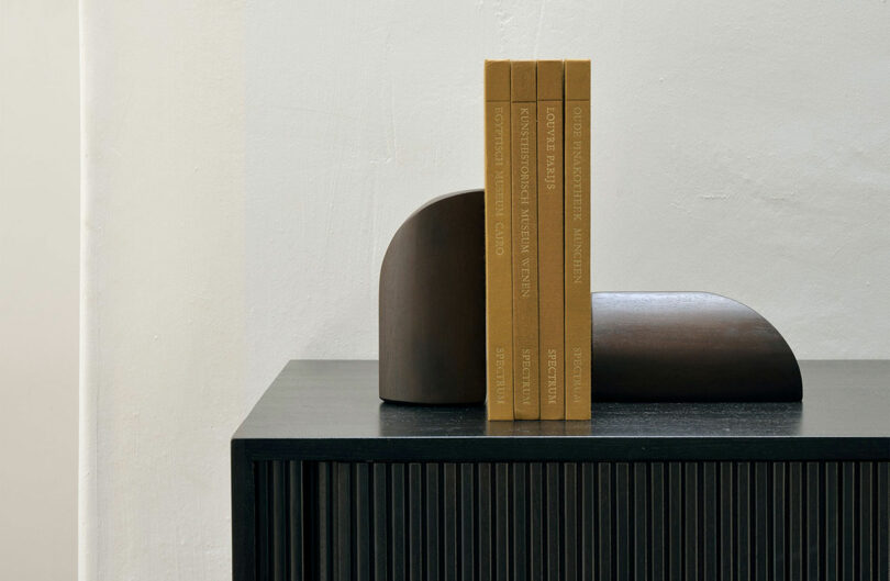 Three books with brown covers stand upright between two curved, wooden bookends on a dark, ribbed cabinet against a plain white wall.