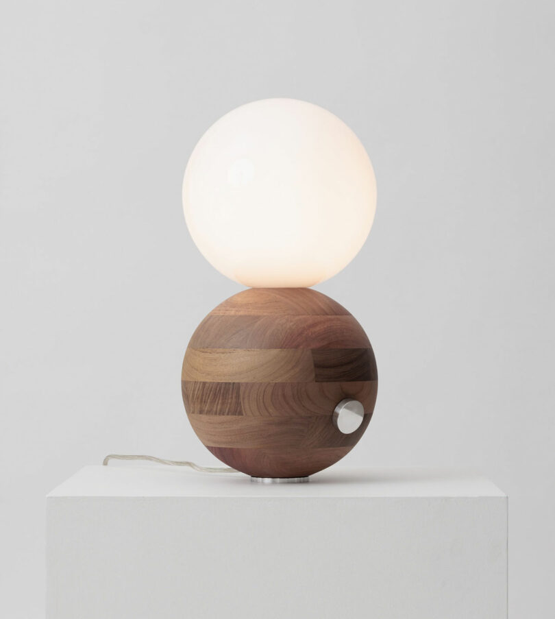 A modern table lamp with a round, glowing white bulb on top of a wooden spherical base, placed on a white pedestal against a neutral background.