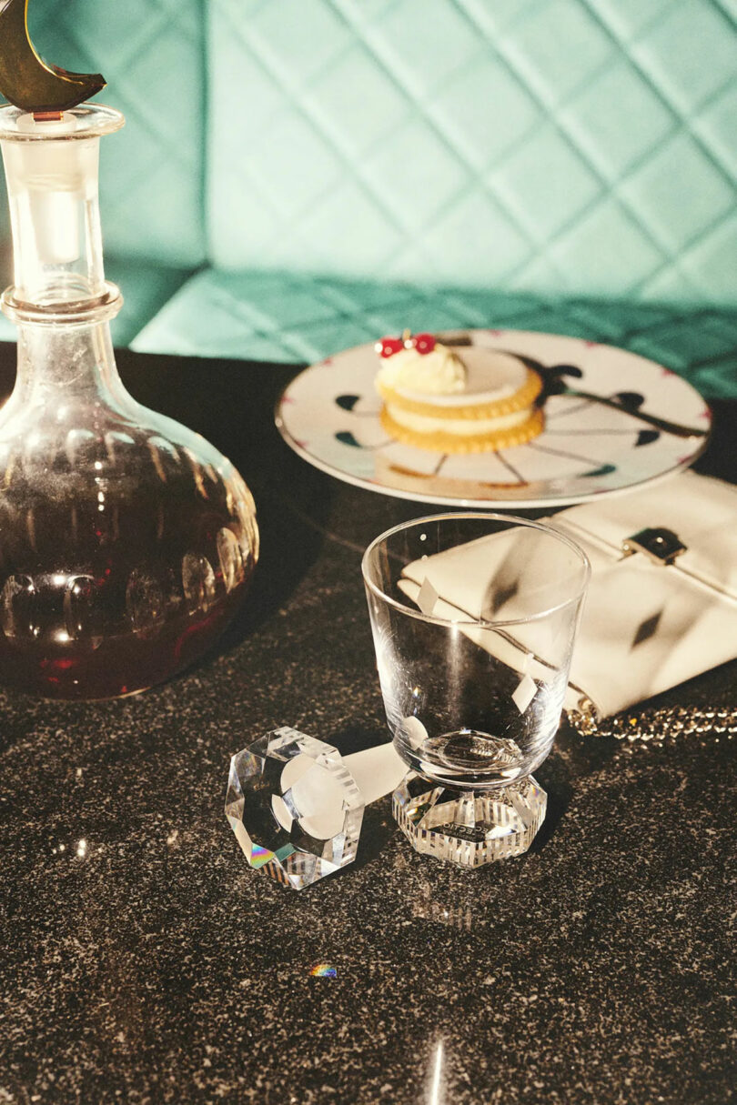 A glass with a crystal stopper lies on a glossy table next to a decanter, showcasing modern gifts. Nearby, a small dessert on a plate accompanies the elegant scene, completed by a chic quilted handbag.