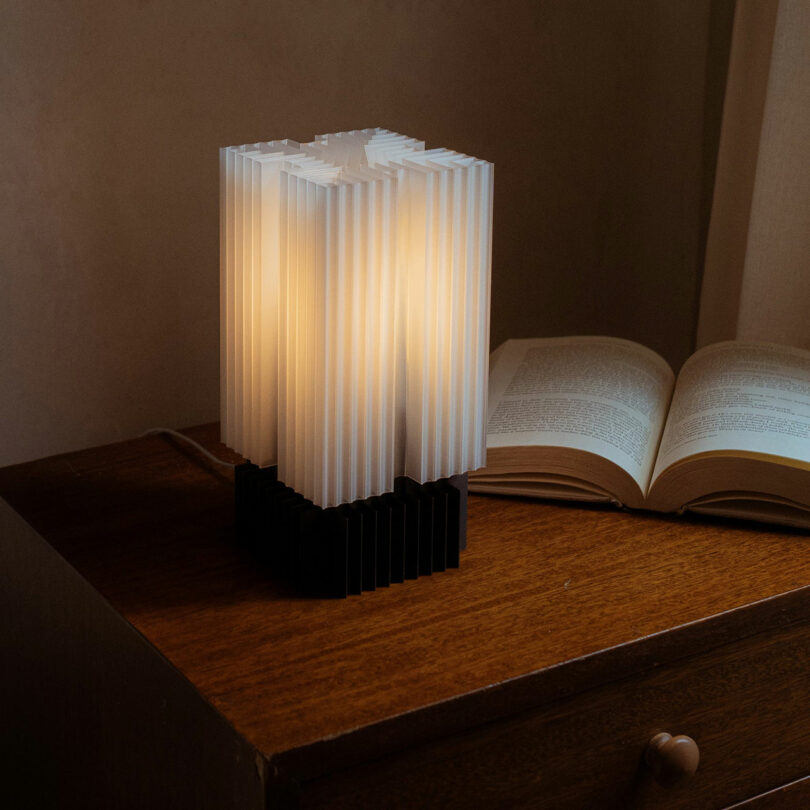 A modern lamp with a ribbed design, perfect for modern gifts, emits warm light on a wooden table, next to an open book.