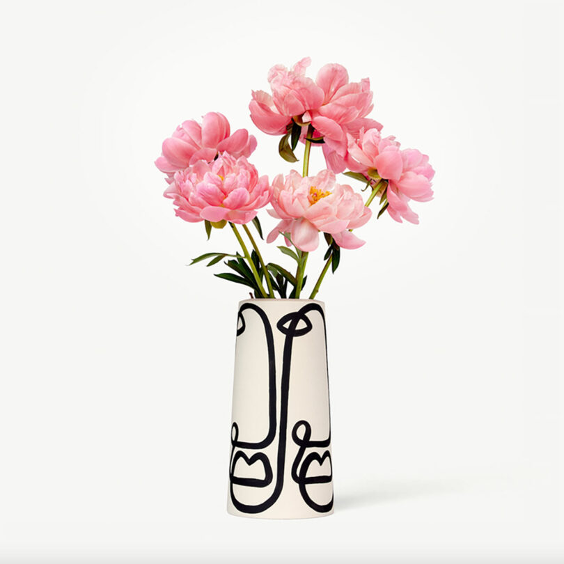 Vase with a black abstract face design, perfect for modern gifts, holding pink peonies against a plain white background.