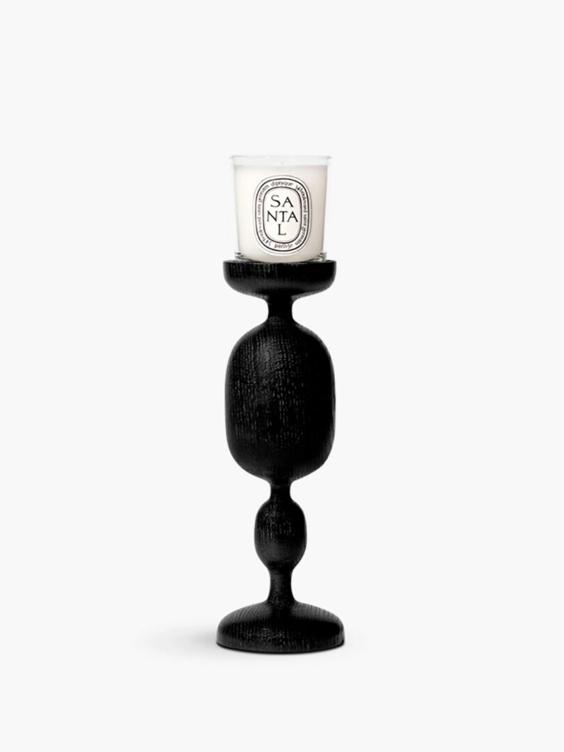 A chic black candlestick holder with an intricately shaped base holds a white "Santal" candle in a glass container, making it the perfect modern gift.