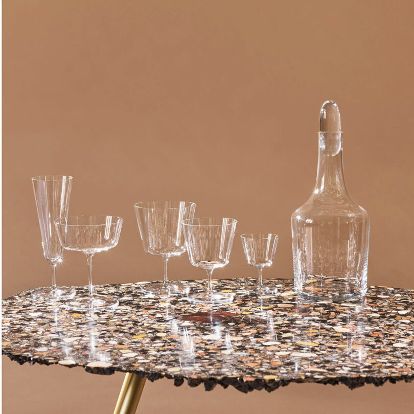 A collection of modern gifts featuring various clear glassware, including a large bottle and several different glasses, displayed on a speckled tabletop against a brown background.