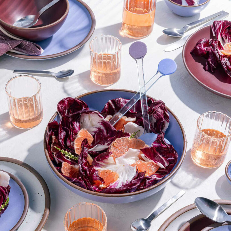 A modern gift to the senses, this salad features red radicchio and orange segments in a blue bowl, surrounded by glasses of pink drink, plates, and cutlery on a white table.