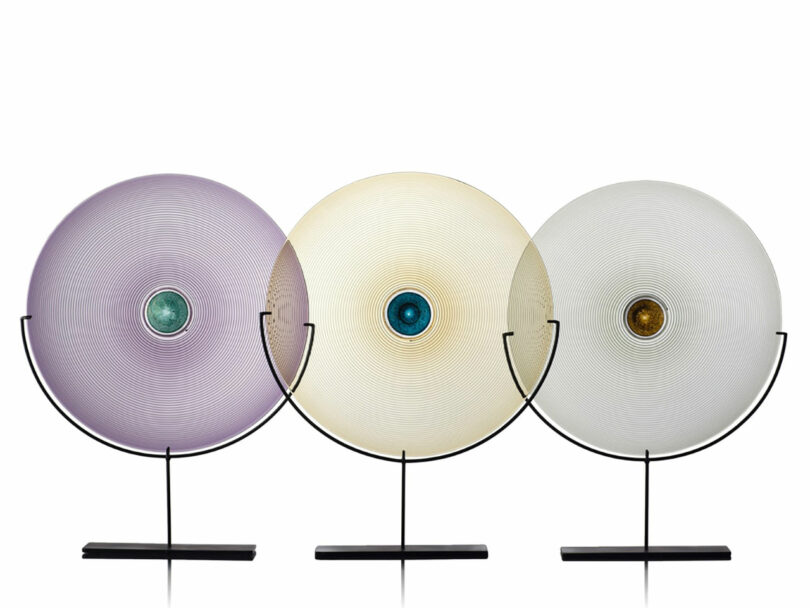 Three circular glass sculptures with solid black bases make stunning modern gifts. The left sculpture is pale purple, the center is beige, and the right is white. Each features a colored center: teal on the left and middle, amber on the right.