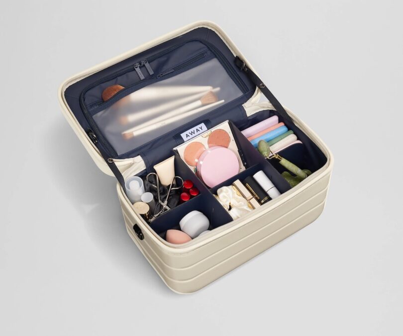 An open beige travel case with multiple compartments, containing makeup brushes, cosmetics, skincare products, and accessories