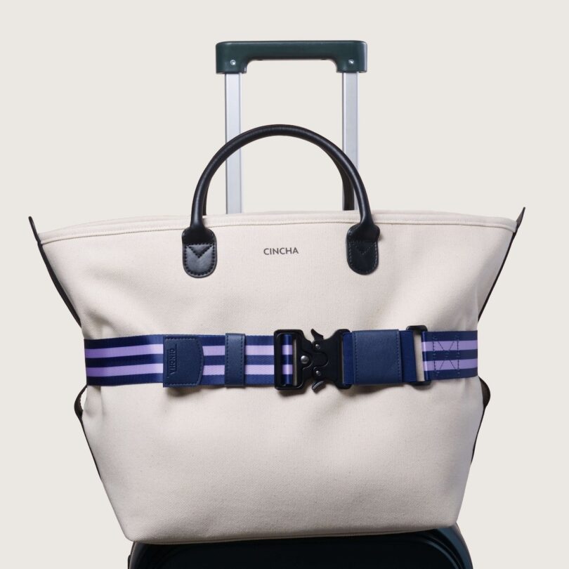 A white travel bag with dark handles is strapped to a rolling suitcase. The strap is blue and purple with a black buckle