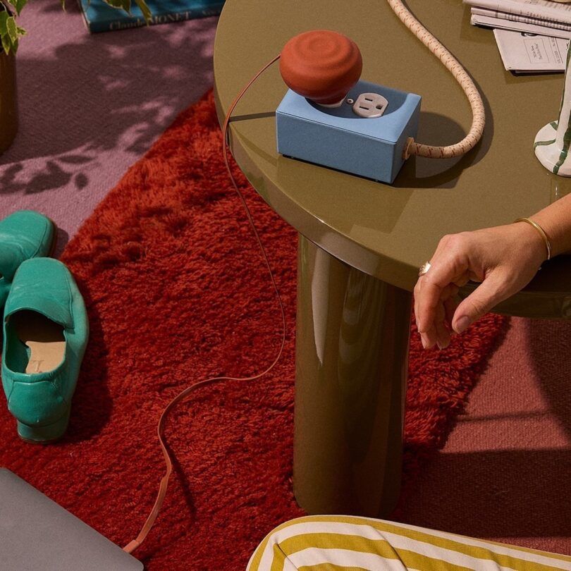 A hand plugs a cord into a blue power outlet on a round table. Nearby are green shoes, a plant, and a red rug