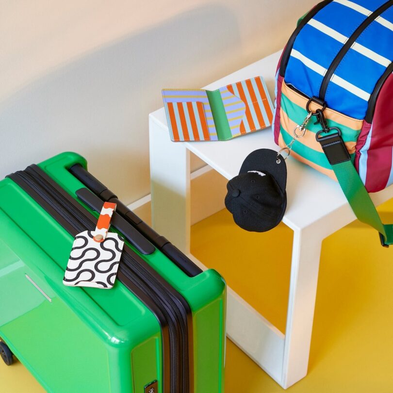 A green suitcase with a patterned luggage tag and a colorful striped bag sit on a white stool. A small black pouch is attached to the bag, and a striped notebook is nearby on the floor