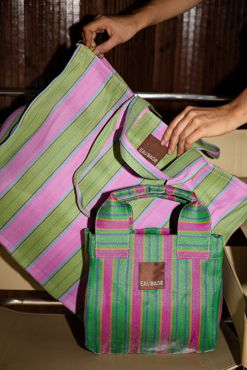 Two striped bags in pink, green, and blue on a chair, with a person's hands holding the larger one. One bag reads "EAU BAGG